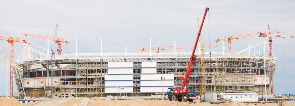 A large stadium facility is under construction, with scaffolding, several cranes, and construction equipment visible on the sandy site in the foreground.