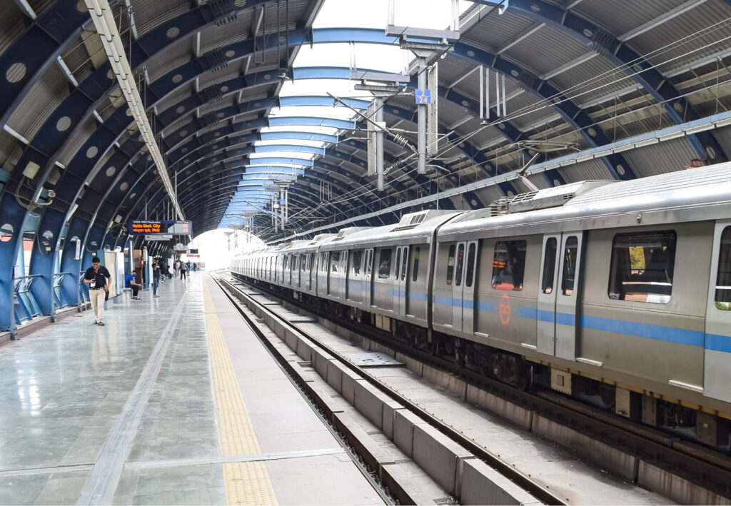 A modern metro train is stopped at an empty platform in a covered urban station, part of a Rail awareness transit campaign, with a few passengers waiting and signs visible overhead.