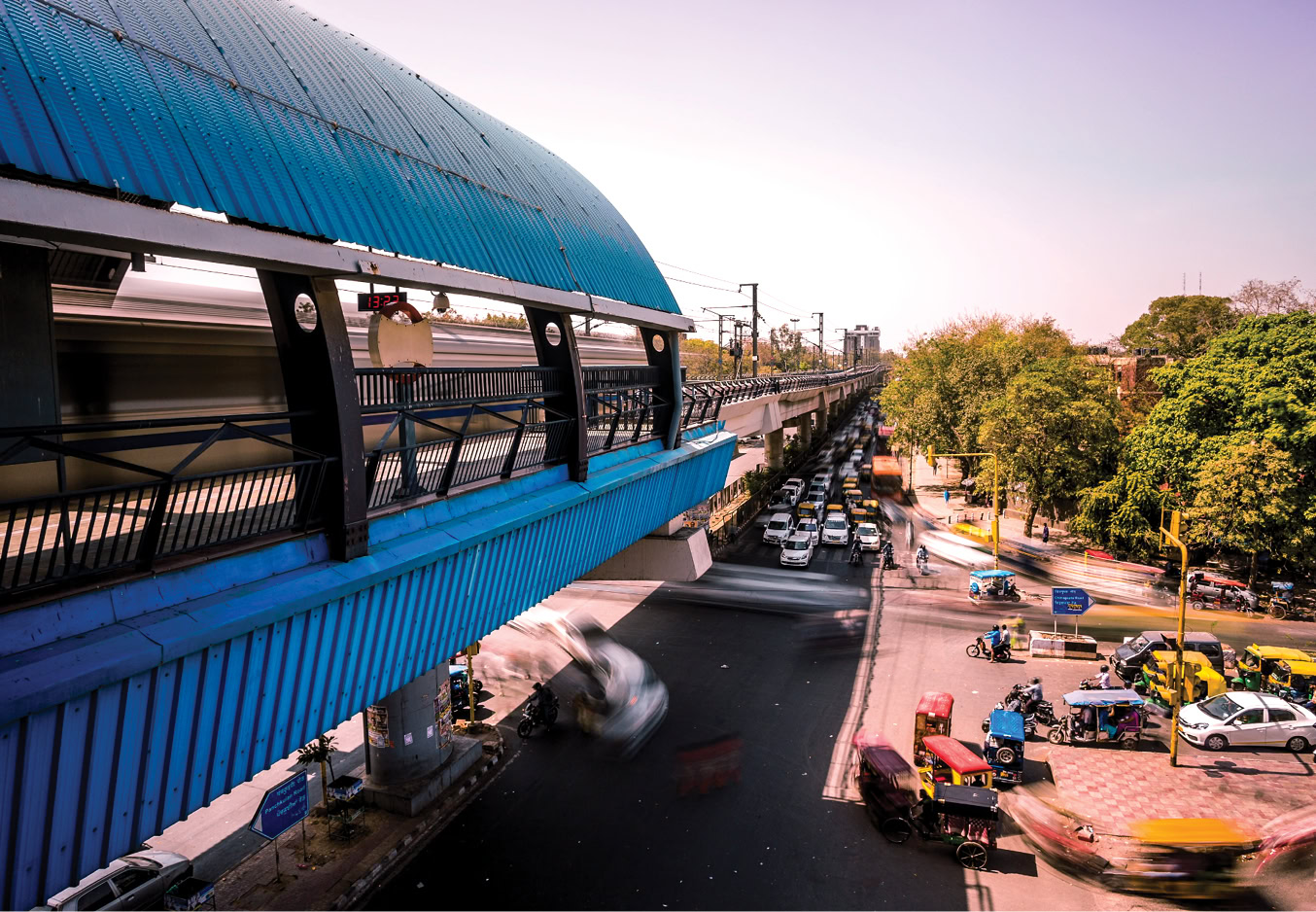 A metro rail train passes through an elevated blue station above a busy road with cars, rickshaws, and trees, under a clear sky.