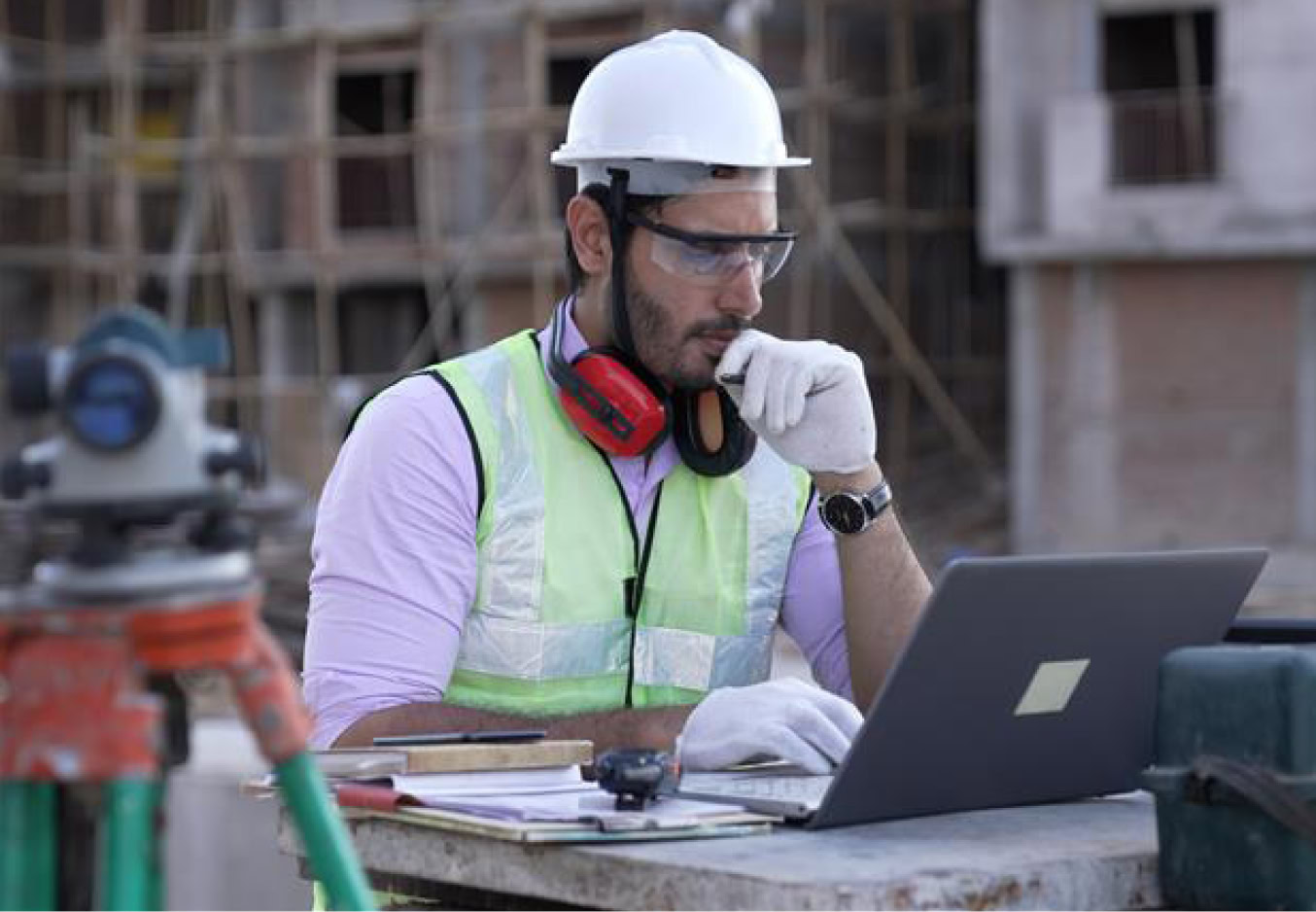 A construction worker in safety gear uses a laptop at an outdoor rail worksite, supporting an awareness campaign near a partially constructed building in the background.