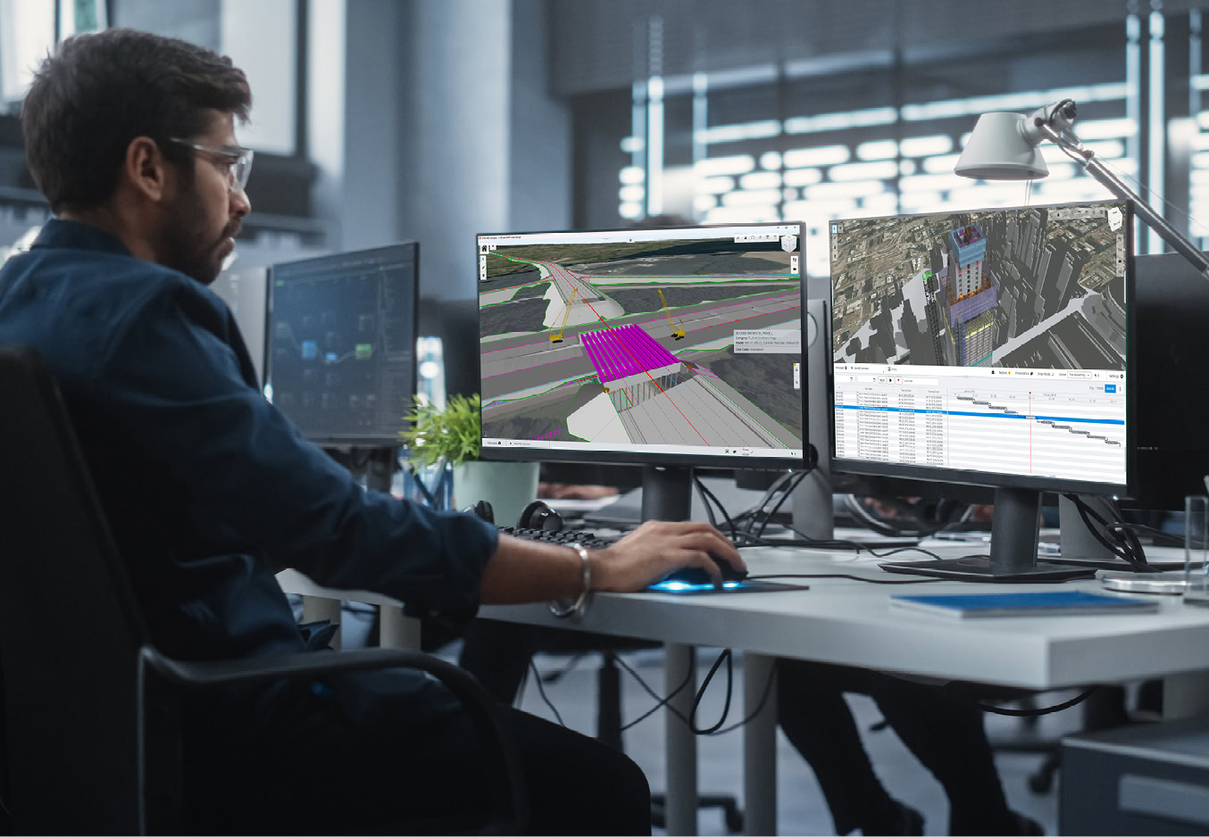 A man sits at a desk using three monitors, reviewing 3D architectural and infrastructure models and a data table in an office setting as part of a rail awareness transit campaign.