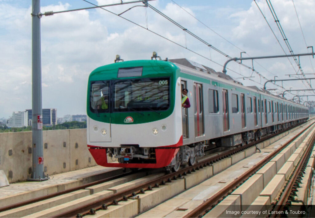 A modern metro rail train travels on an elevated track under an overhead catenary system, with drivers visible in the front cab, highlighting efficient urban transit.