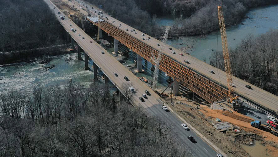 Aerial view of a highway bridge under construction next to an existing bridge over a river, with cranes and vehicles present.