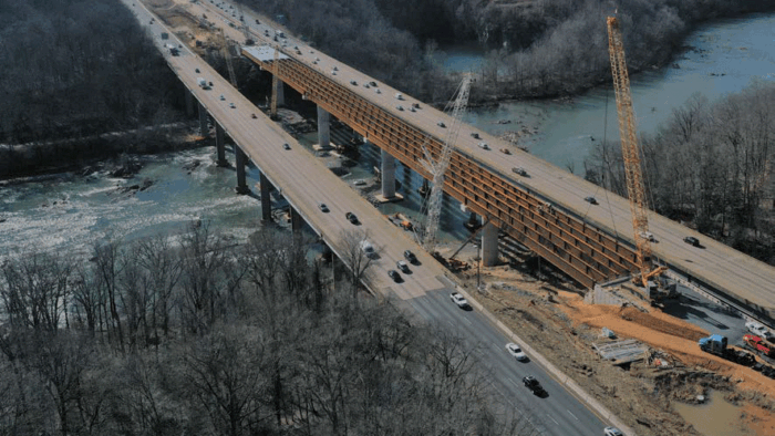Aerial view of a highway bridge under construction beside an existing bridge over a river, with cranes and vehicles present—showcasing advanced transportation solutions from Bentley Systems.