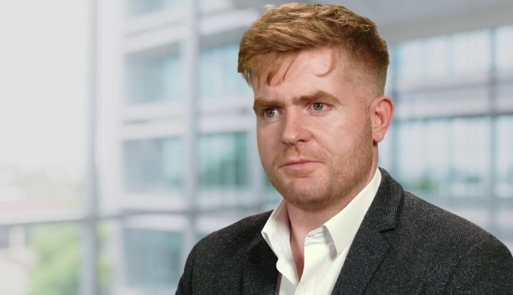 A man with short light brown hair wearing a white shirt and dark blazer sits indoors in front of large, blurred windows, reflecting on his CAD Journey as a Partner of the Year.