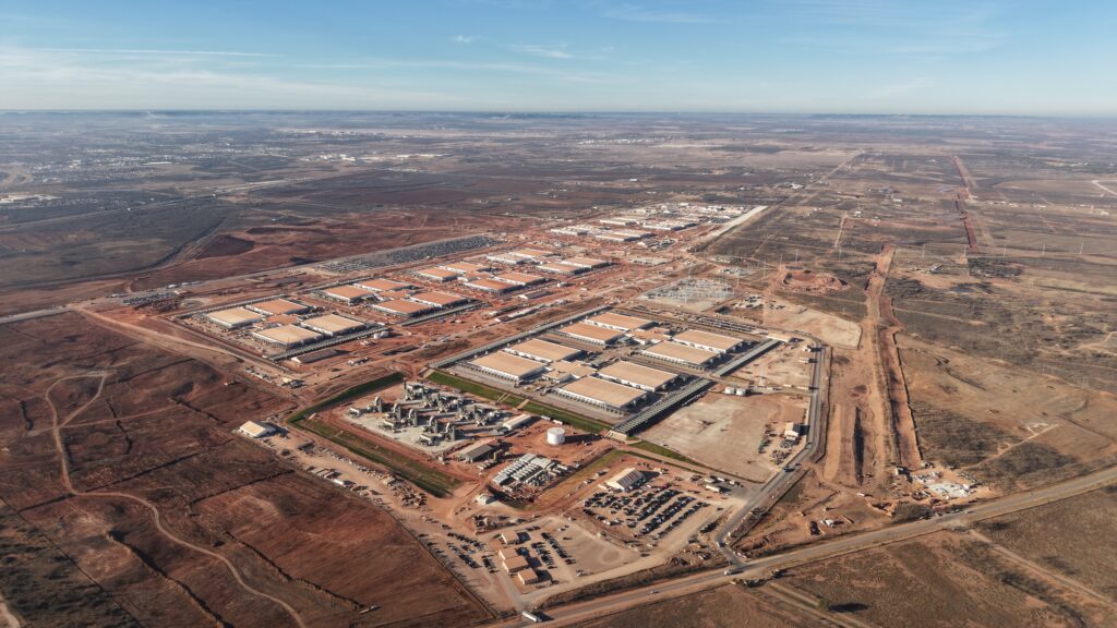 Aerial view of a large industrial complex featuring multiple data centers, construction areas, and parking lots, surrounded by barren land and roads.