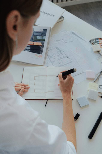 Person sketching architectural plans in a notebook at a desk with building blueprints, samples, and a book titled "Bauhaus," illustrating the Engineering Design Process in action.