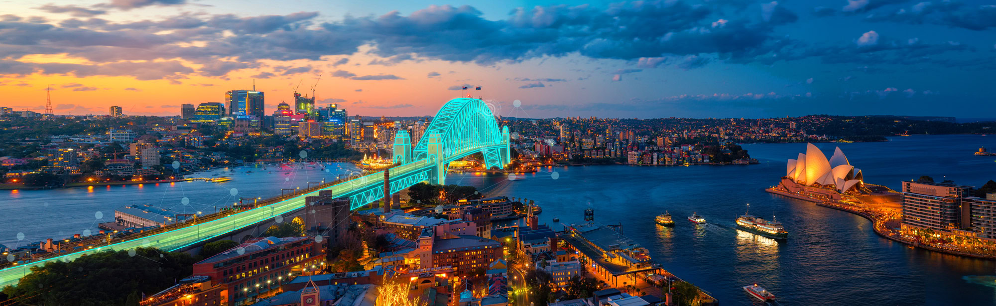 A panoramic view of Sydney at twilight, featuring the illuminated Sydney Harbour Bridge, the Sydney Opera House, and the city skyline with buildings lit up. Downloading this image is like saying thank you to your senses for such a breathtaking experience.