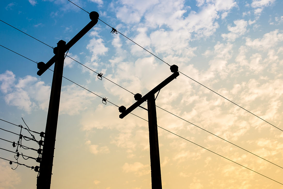 Silhouettes of utility poles and power lines, essential to electric utilities, stand against a sky with scattered clouds at sunset or sunrise.