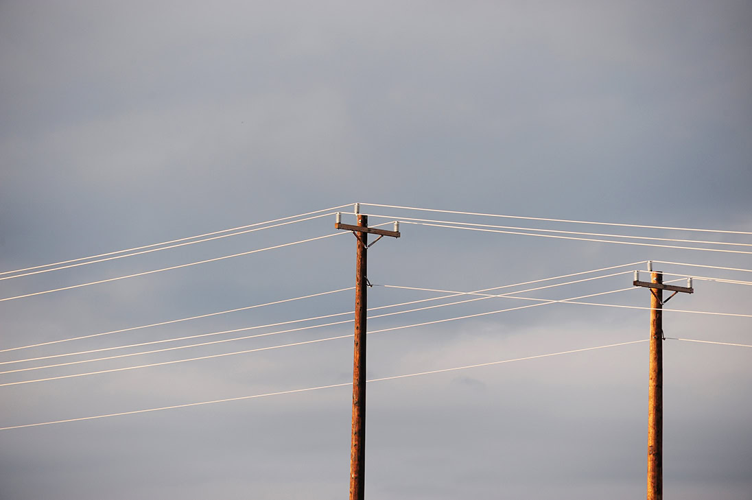 Two utility poles with multiple electric power lines stretch across a cloudy sky, the poles and wires silhouetted against the light background.