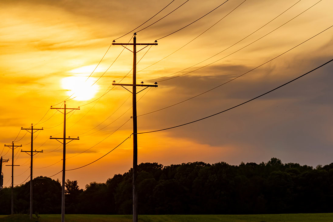 Power lines supporting electric utilities stretch across wooden utility poles in a field at sunset, framed by trees and an orange sky.