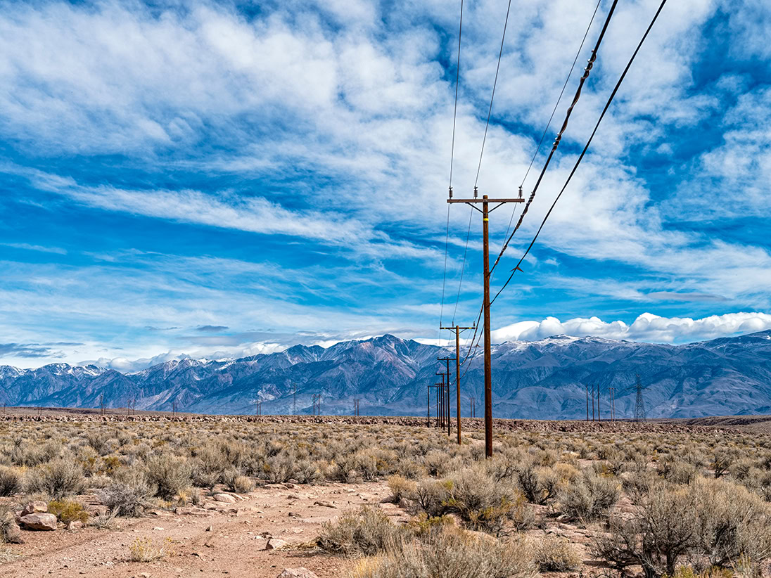 Utility poles with power lines from local electricity providers stretch across a dry, scrubland desert landscape, set against distant mountains and a clear blue sky.