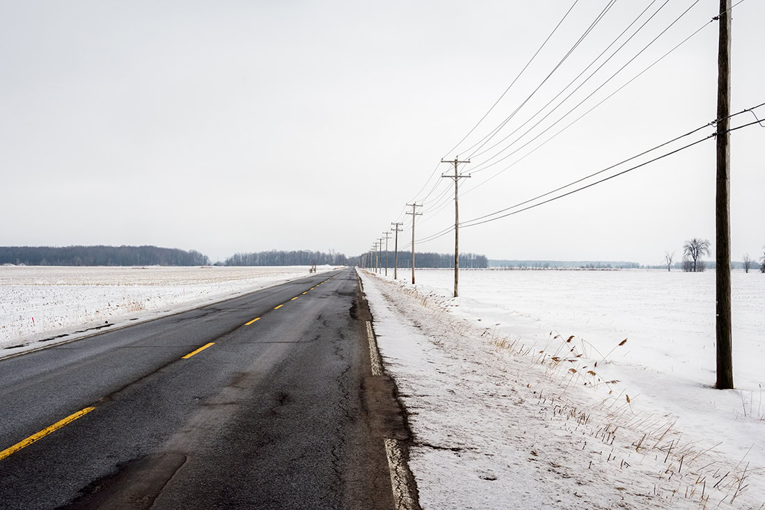 A two-lane road runs through a flat, snow-covered landscape with electric utilities and utility poles lining the right side under an overcast sky.