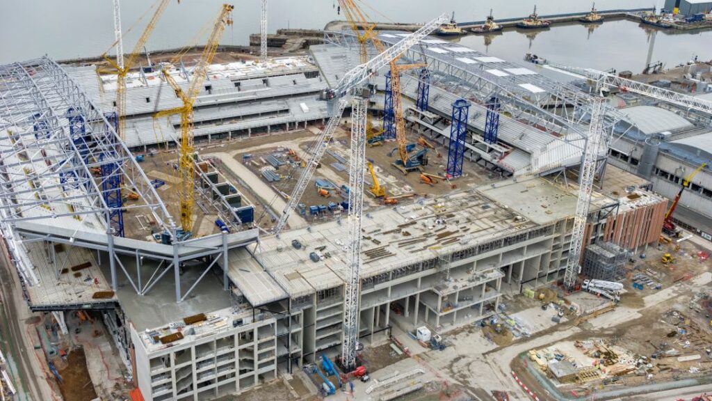 Aerial view of a large construction site with cranes, steel structures, and partially built buildings near a waterfront, perfectly illustrating the complexity and efficiency showcased in this SYNCHRO Case Study.