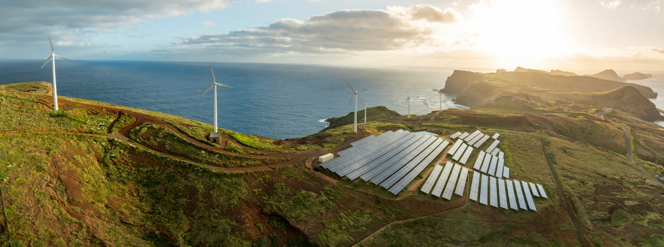 Wind turbines and solar panels are installed on a grassy coastal hillside overlooking the ocean under a partly cloudy sky at sunset, demonstrating real-world applications perfect for electrical training or EasyPower electrical courses.