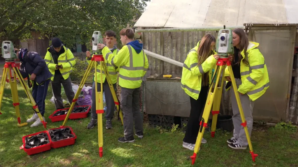 Five people in high-visibility jackets use surveying equipment on tripods in a grassy outdoor area near a greenhouse, collaborating on a Design Challenge focused on achieving Zero Carbon solutions.
