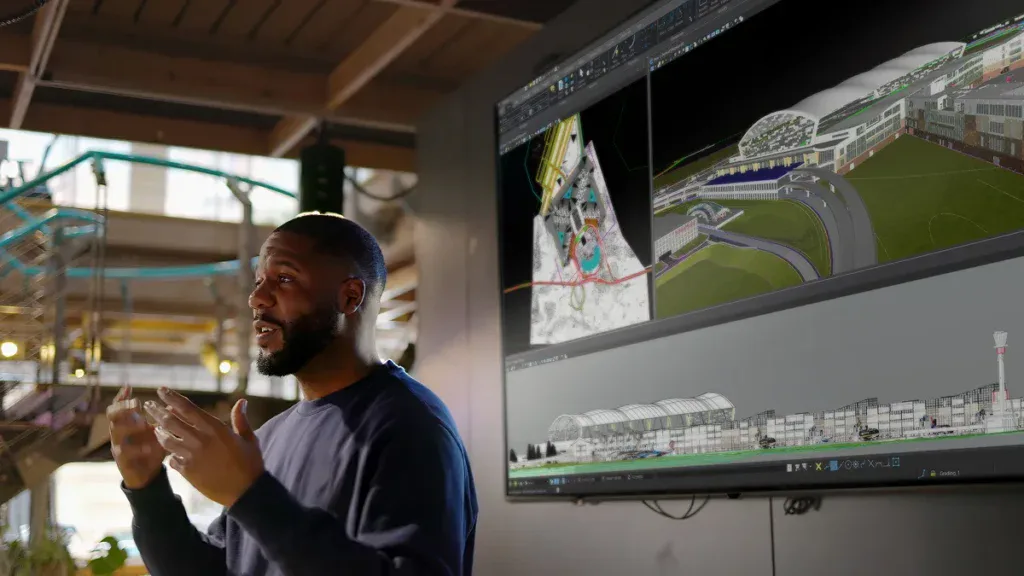 A man gestures while giving a presentation beside a large screen displaying architectural and 3D building models.