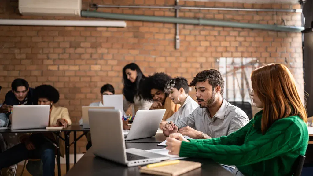 A group of people sit at tables in a modern classroom, working on laptops and writing notes with a brick wall in the background.