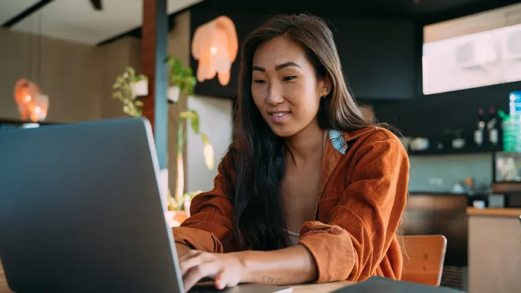 A woman sits at a table using a laptop in a modern kitchen with plants and warm lighting in the background.