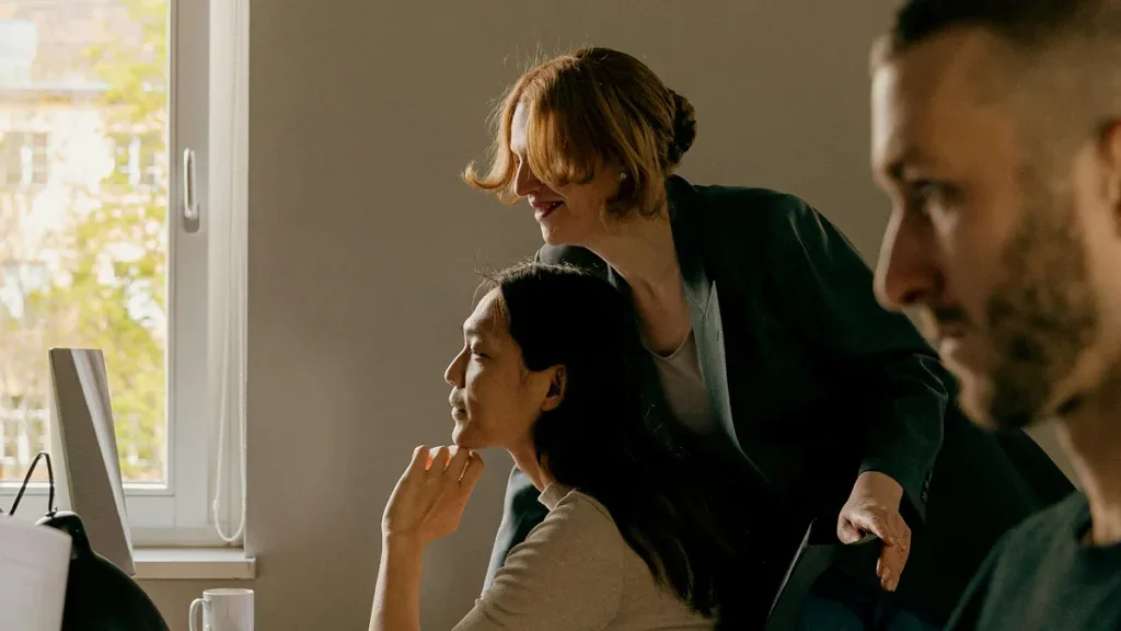 Three people are working in an office; two women look at a computer screen together while a man sits in the foreground.