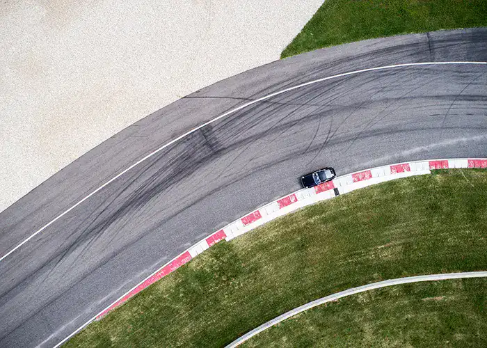 Aerial view of a car driving on a curved racetrack, with tire marks visible on the asphalt and grass bordering the track.