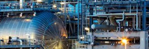 An industrial facility with a complex network of pipelines, tanks, and structures illuminated by lights at dusk.