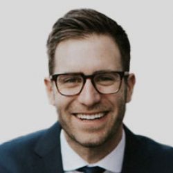 A man with short brown hair and glasses is smiling, wearing a dark suit, white shirt, and tie, against a plain light background—reflecting confidence in the field of Bridge Information Modeling (BIM) and AI solutions.