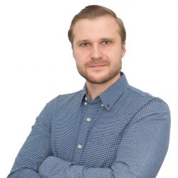 A person with light hair and a beard is wearing a blue checkered shirt and standing with arms crossed against a white background.