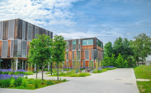 Modern buildings with wooden accents stand amid lush greenery and blooming flowers under a blue sky with scattered clouds.