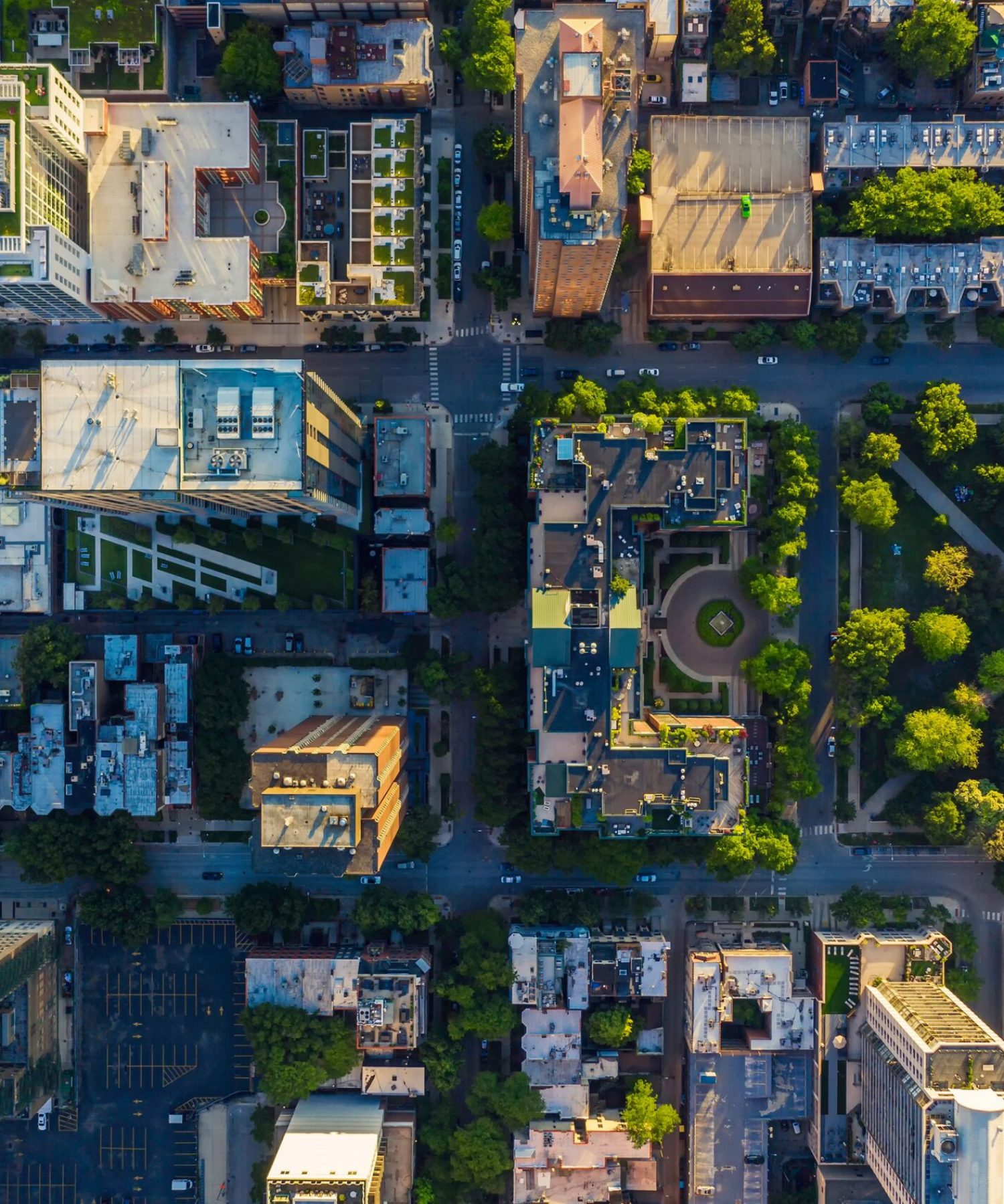 Aerial view of a city block with buildings, roads, and green park areas arranged in a grid layout, captured in daylight.