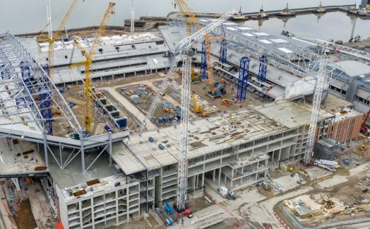 Aerial view of a large construction site with cranes, steel structures, and partially built buildings near a waterfront, perfectly illustrating the complexity and efficiency showcased in this SYNCHRO Case Study.