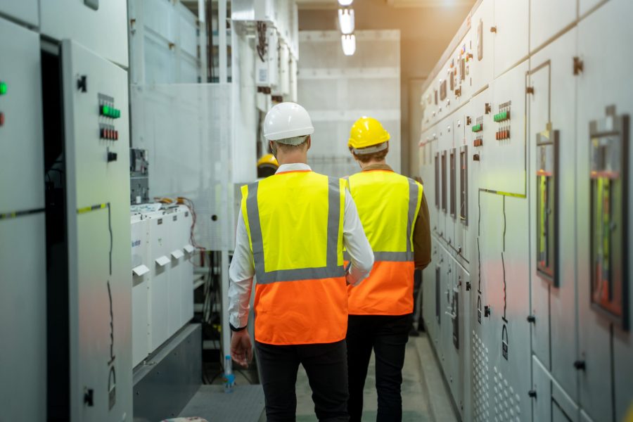 Two workers wearing safety vests and helmets walk through an industrial control room lined with electrical panels, demonstrating the importance of electrical training in real-world settings.