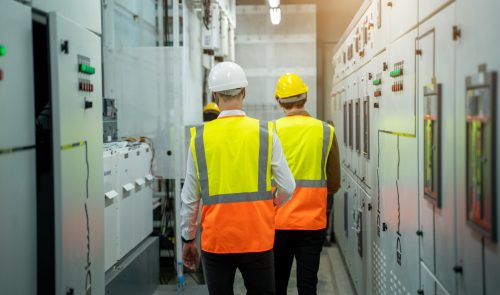Two workers wearing safety vests and helmets walk through an industrial control room lined with electrical panels, demonstrating the importance of electrical training in real-world settings.