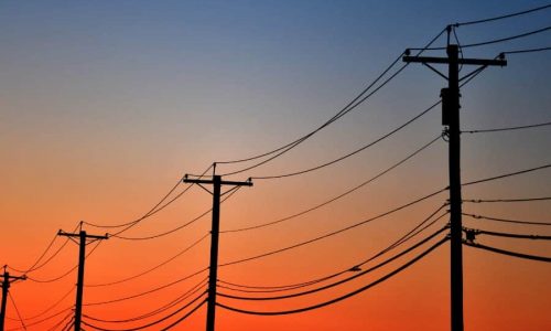 Multiple utility poles with power lines silhouetted against an orange and blue gradient sky at sunset.