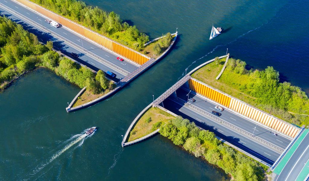 Aerial view of a canal bridge with cars on roads above and boats navigating the water below. Greenery surrounds the bridge.