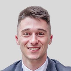 Young man with short brown hair, wearing a gray suit jacket and white shirt, smiling at the camera against a plain light gray background, representing the next generation of BIM and AI professionals.