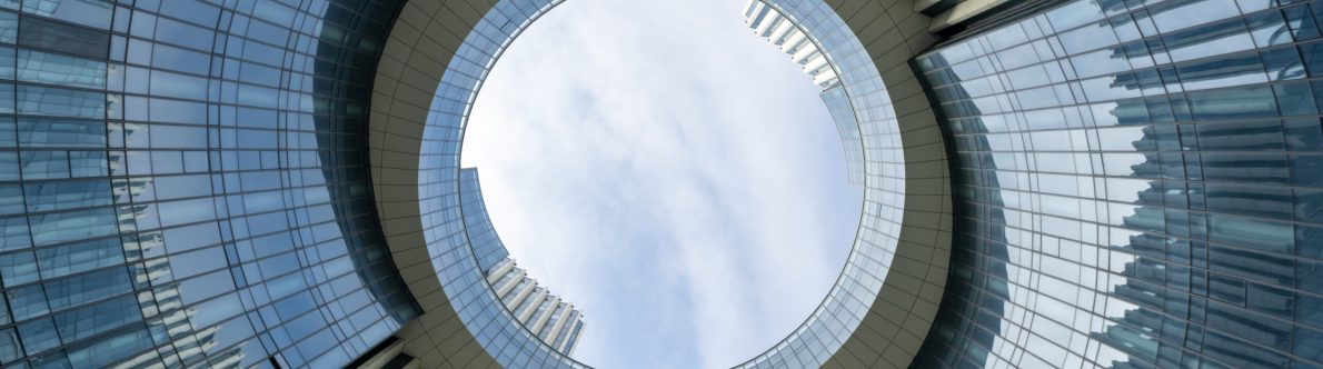 Upward view of two modern glass buildings connected by a large circular skybridge with an open center, reflecting the innovation of open infrastructure, set against a partly cloudy sky.