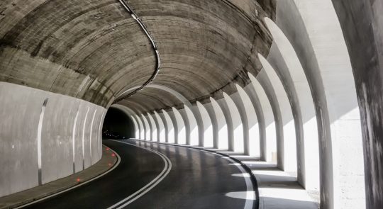 Curved road tunnel with concrete arches and white dividing lines, featuring light and shadow patterns on the walls and pavement—a true architectural spotlight.
