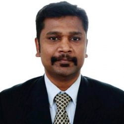 A man with a mustache wearing a black suit, white shirt, and patterned tie against a plain white background.