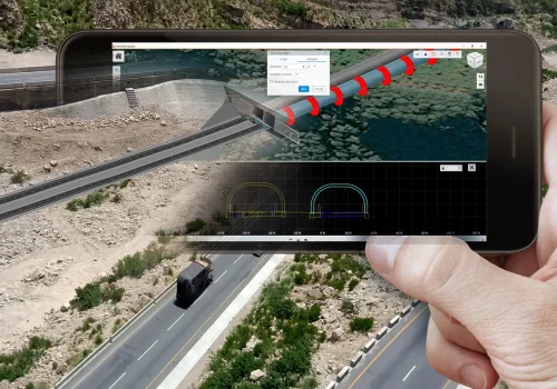 A person's hand holding a smartphone with the SYNCHRO Digital Construction Software, displaying a 3D modeling app that overlays a digital barrier on a real roadway beside a river.