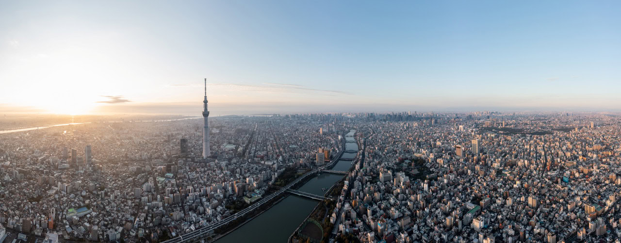 A wide aerial view of Tokyo at sunrise, showing dense city buildings, the Tokyo Skytree tower, and a river running through the city.