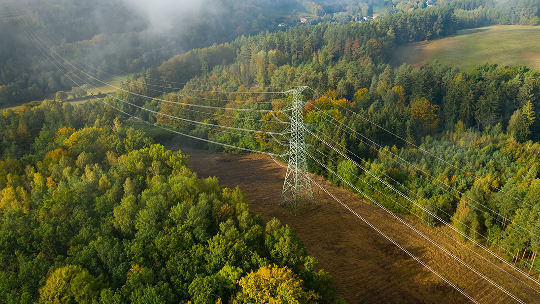 Aerial view of a metal electricity transmission tower and power lines, part of the electric utilities network, running through a forested landscape with trees and grassy clearings.