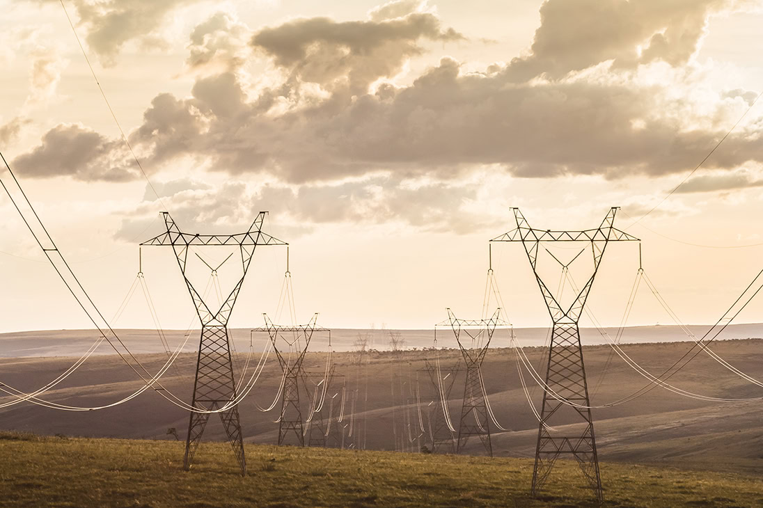 High-voltage power lines supported by metal pylons, part of vital electric utilities infrastructure, stretch across an open landscape under a cloudy sky at sunset or sunrise.