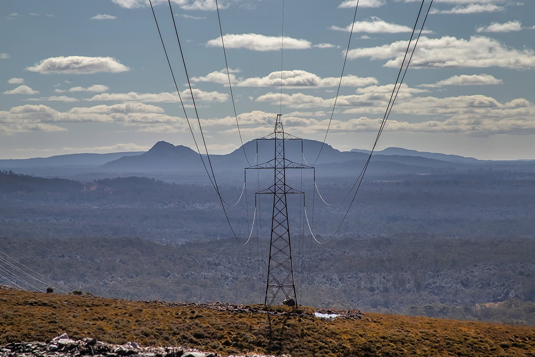 A metal electricity pylon stands in open terrain, a striking symbol of electric utilities, with power lines stretching into the distance; hills and a partly cloudy sky form the backdrop.