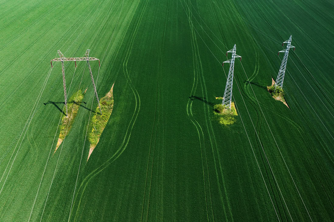Aerial view of two sets of electric utility towers in a green field, with visible vehicle tracks and patches of exposed soil around the bases.