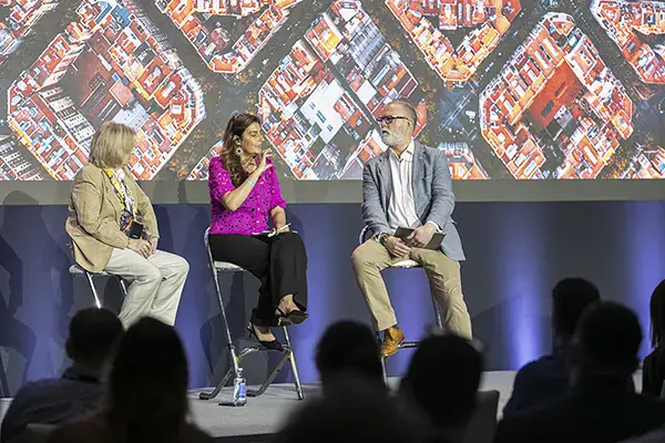 Three people sit on stage having a discussion in front of an audience, with an aerial cityscape image projected behind them.