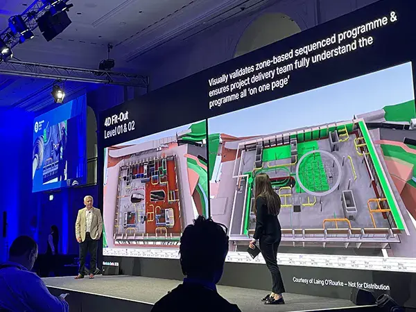 A man presents in front of a large screen displaying 3D building plans and text about validating zone-based sequenced project plans at a conference or seminar.
