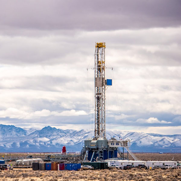 A drilling rig operates in a dry, open landscape with mountains and cloudy skies in the background.