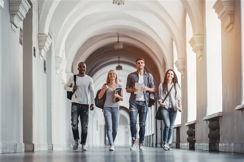 Four Gen Z young adults walk down a bright hallway with arched ceilings, casually dressed and smiling, carrying bags and notebooks—reflecting shared values and vision for the future.