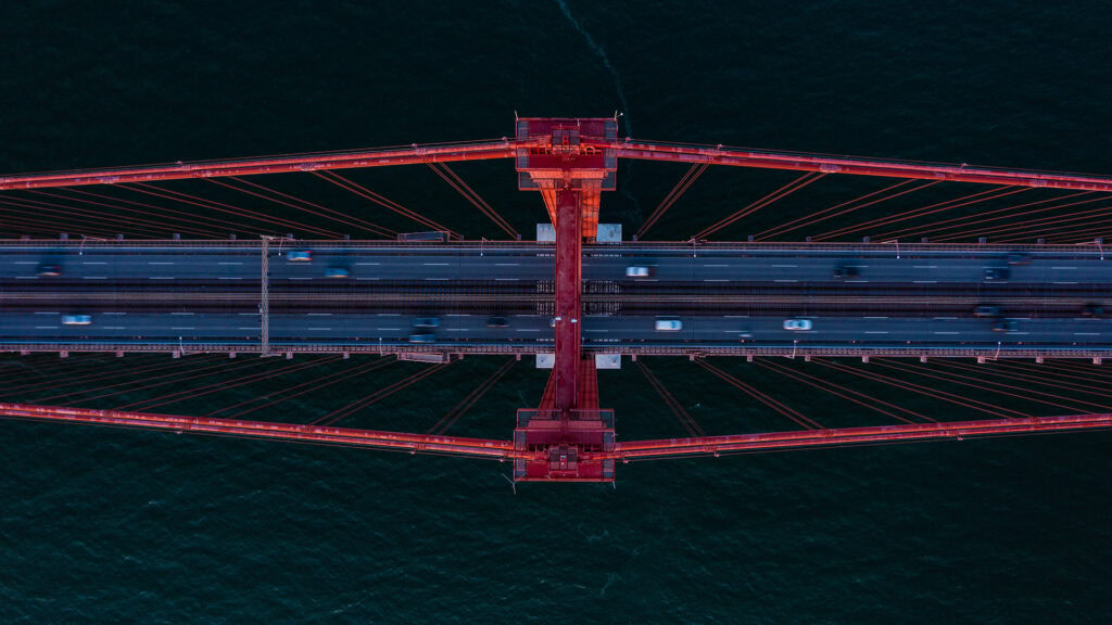 Aerial view of vehicles crossing a large red suspension bridge over dark water.
